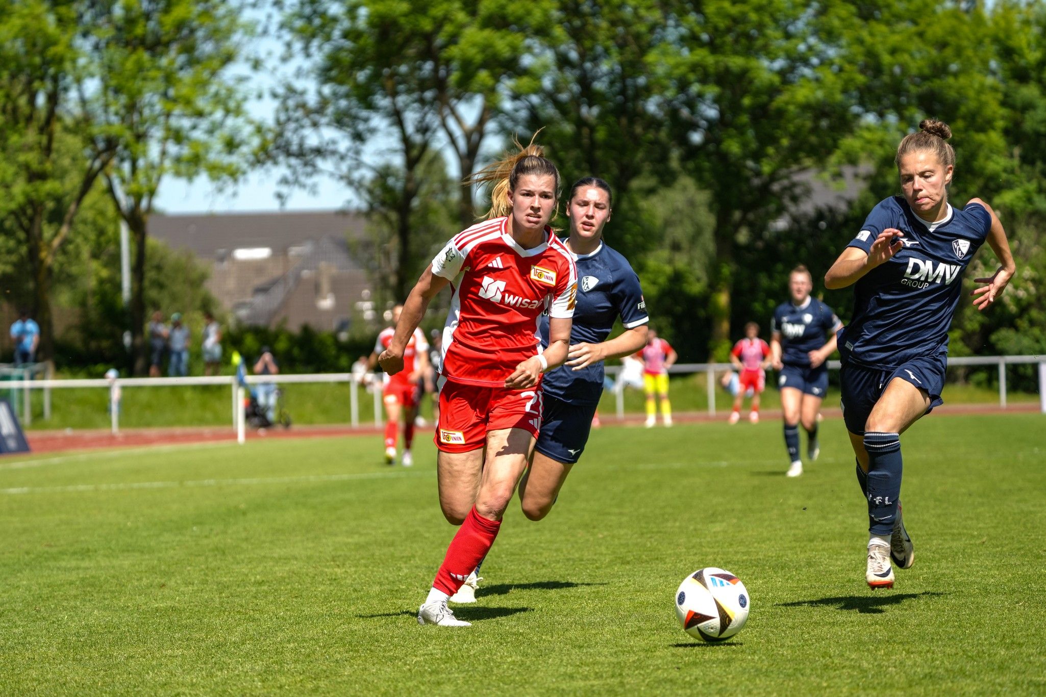 Dos futbolistas con camisetas rojas y azules corren en un campo de hierba, mientras una jugadora dribla el balón.