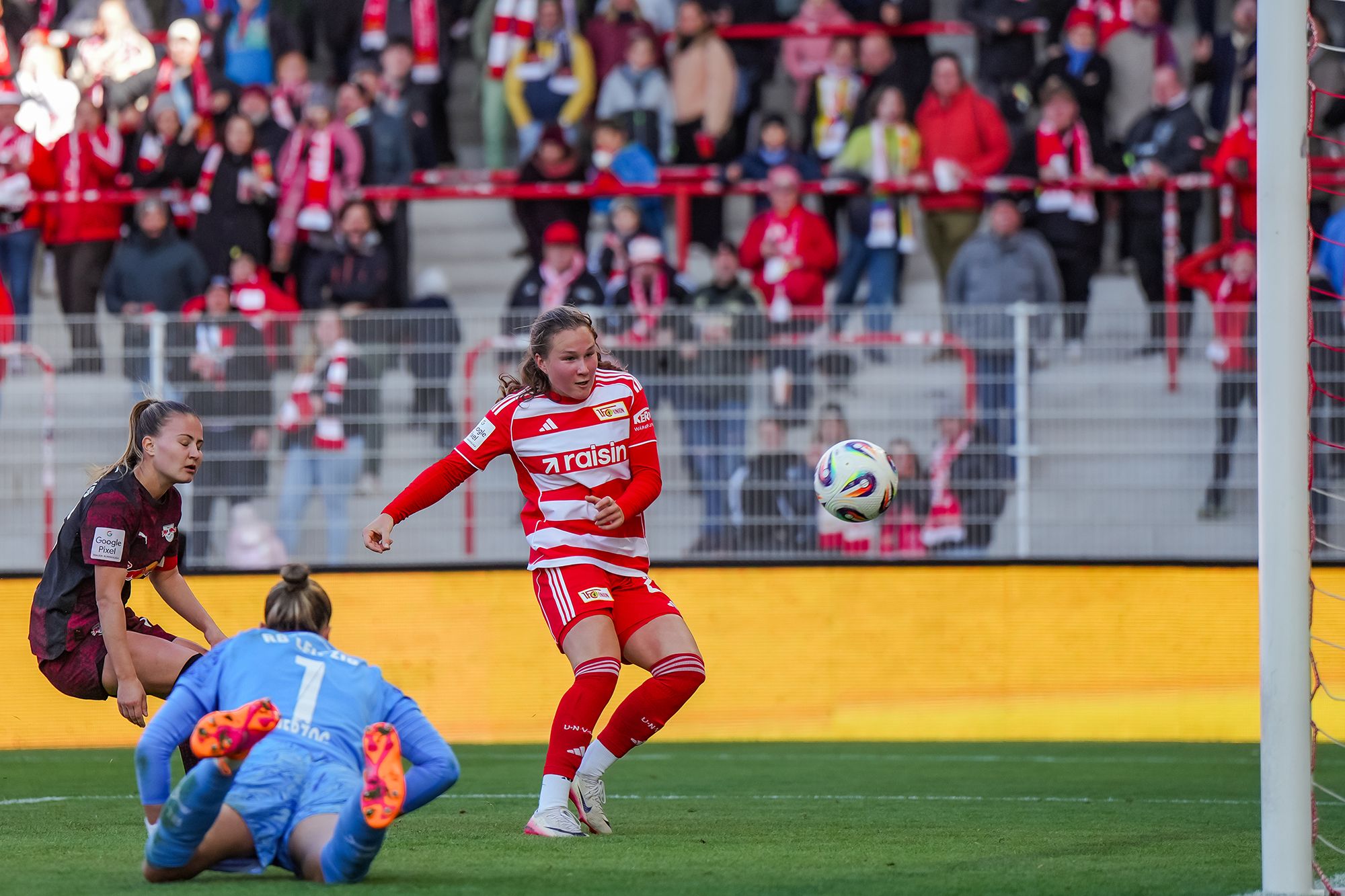 Eine Spielerin in rotem Trikot schießt auf das Tor, während die Torhüterin in blau nach dem Ball greift. Zuschauer im Hintergrund.