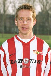 Ein Fußballspieler in einem rot-weißen Trikot mit dem Logo eines Vereins steht vor einem unscharfen Sportplatz.