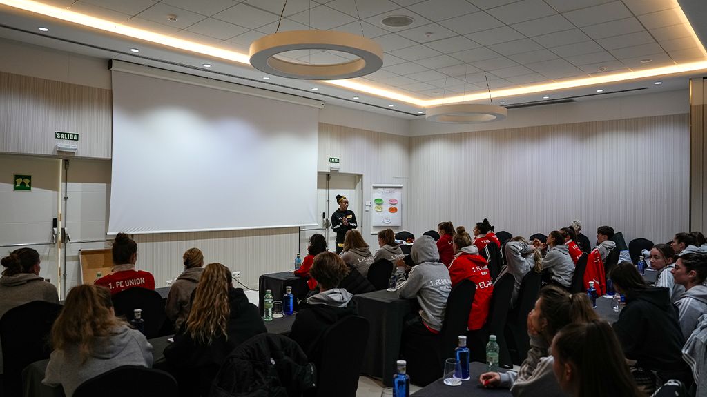 Eine Gruppe von Frauen in roten Sweatshirts sitzt in einem Konferenzraum und hört einer Person vor einem Projektor zu.