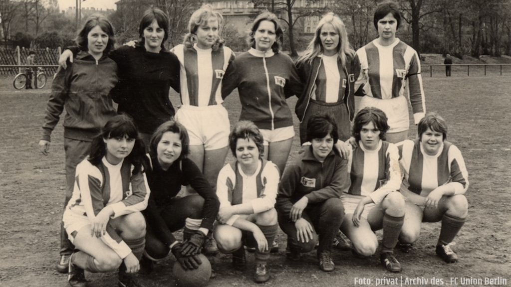Eine Gruppe von Frauen in Fußballtrikots steht auf einem Sportplatz, zusammen mit einem Ball, teils lächelnd, teils ernst.