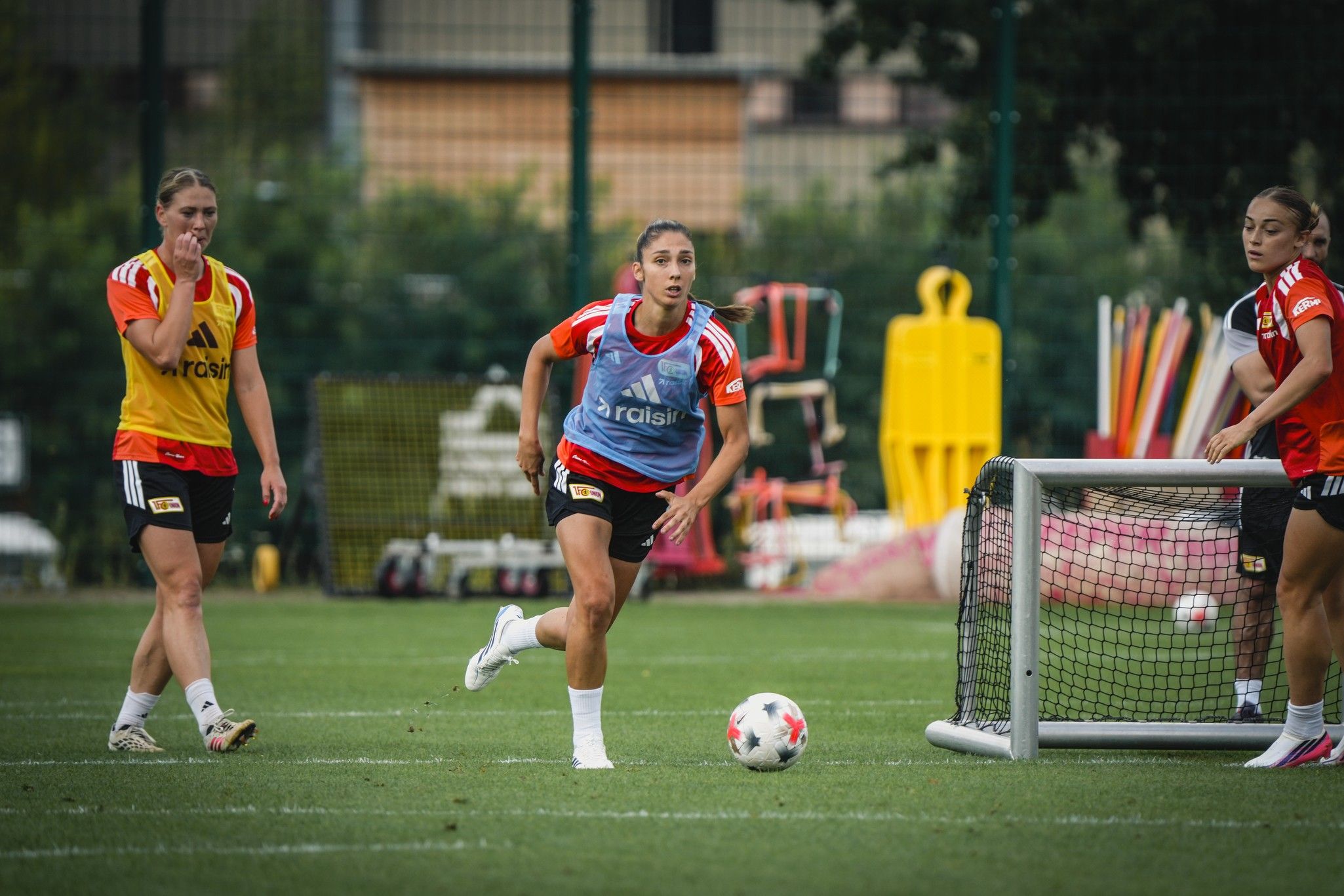 Drei Frauen in Sportkleidung trainieren auf einem Fußballplatz. Eine Spielerin dribbelt den Ball, während die anderen zuschauen.