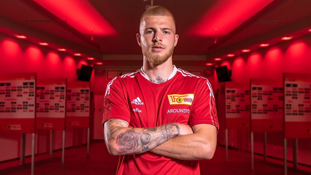 A young man with a beard and tattoos wearing a red soccer jersey stands in front of a bright red wall.