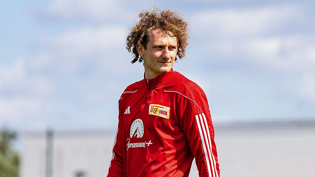 A soccer player with curly hair is wearing a red jersey and gazing thoughtfully into the distance.