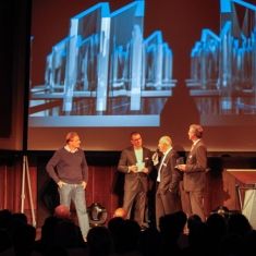 Vier Männer stehen auf einer Bühne vor einer großen Bildschirmpräsentation mit Glasobjekten im Hintergrund.