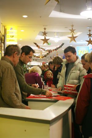 Menschen stehen in einer Halle, während ein Mann Tische bedient und mehrere Personen ihm zuschauen, Weihnachtsdekoration im Hintergrund.