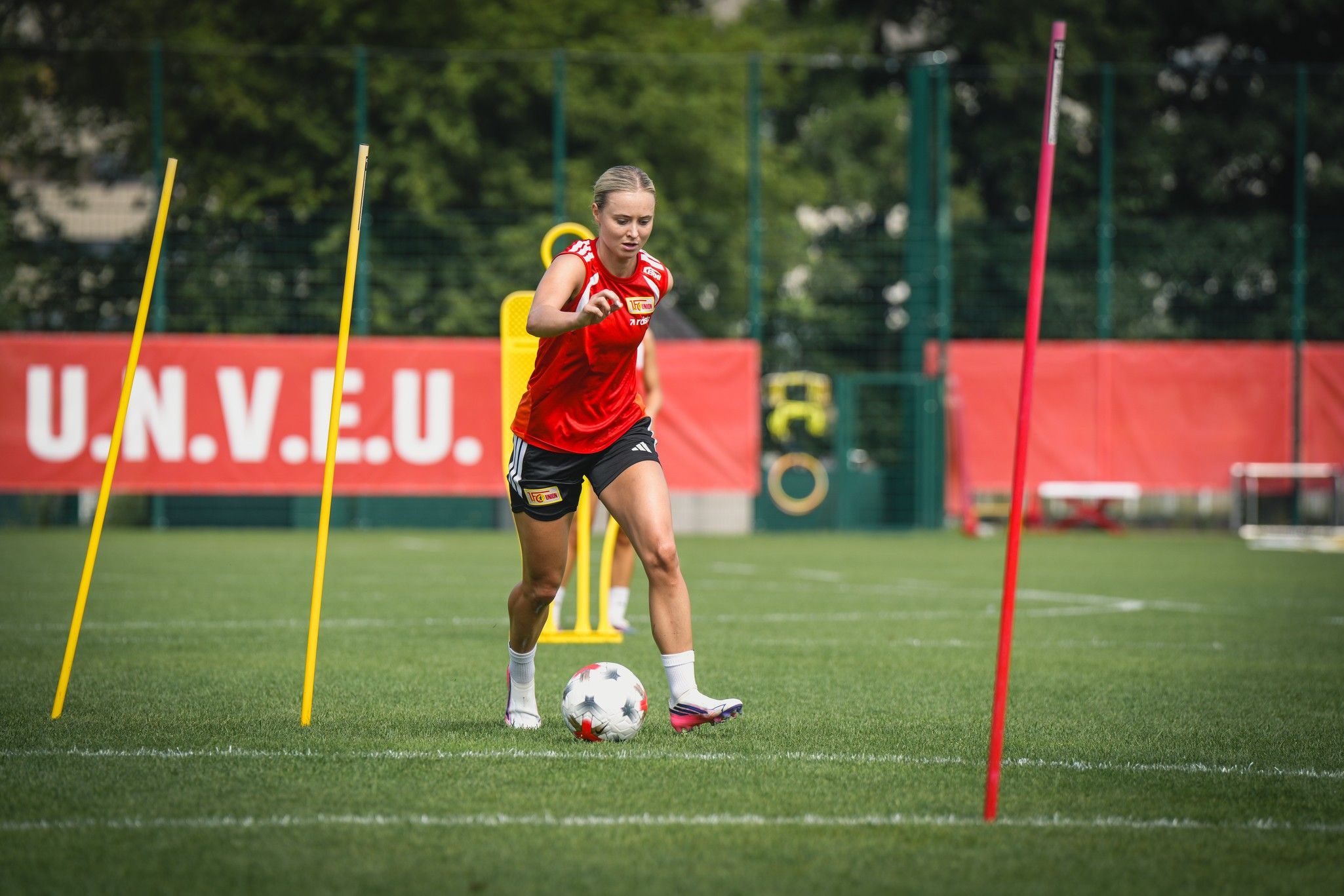 Eine Fußballspielerin in rotem Trikot dribbelt einen Ball im Training und umläuft Hütchen auf einem grünen Sportplatz.