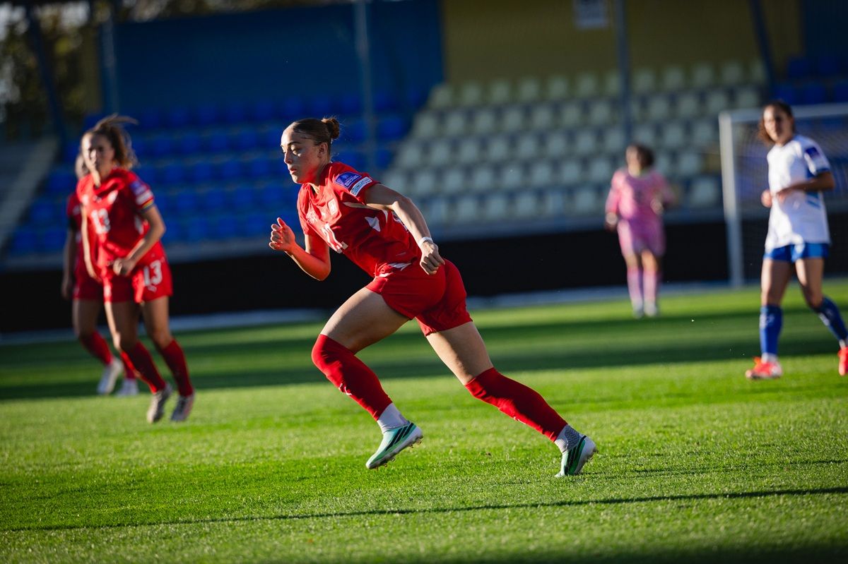 Eine weibliche Fußballspielerin in einem roten Trikot läuft über ein grünes Spielfeld, während andere Spielerinnen im Hintergrund stehen.