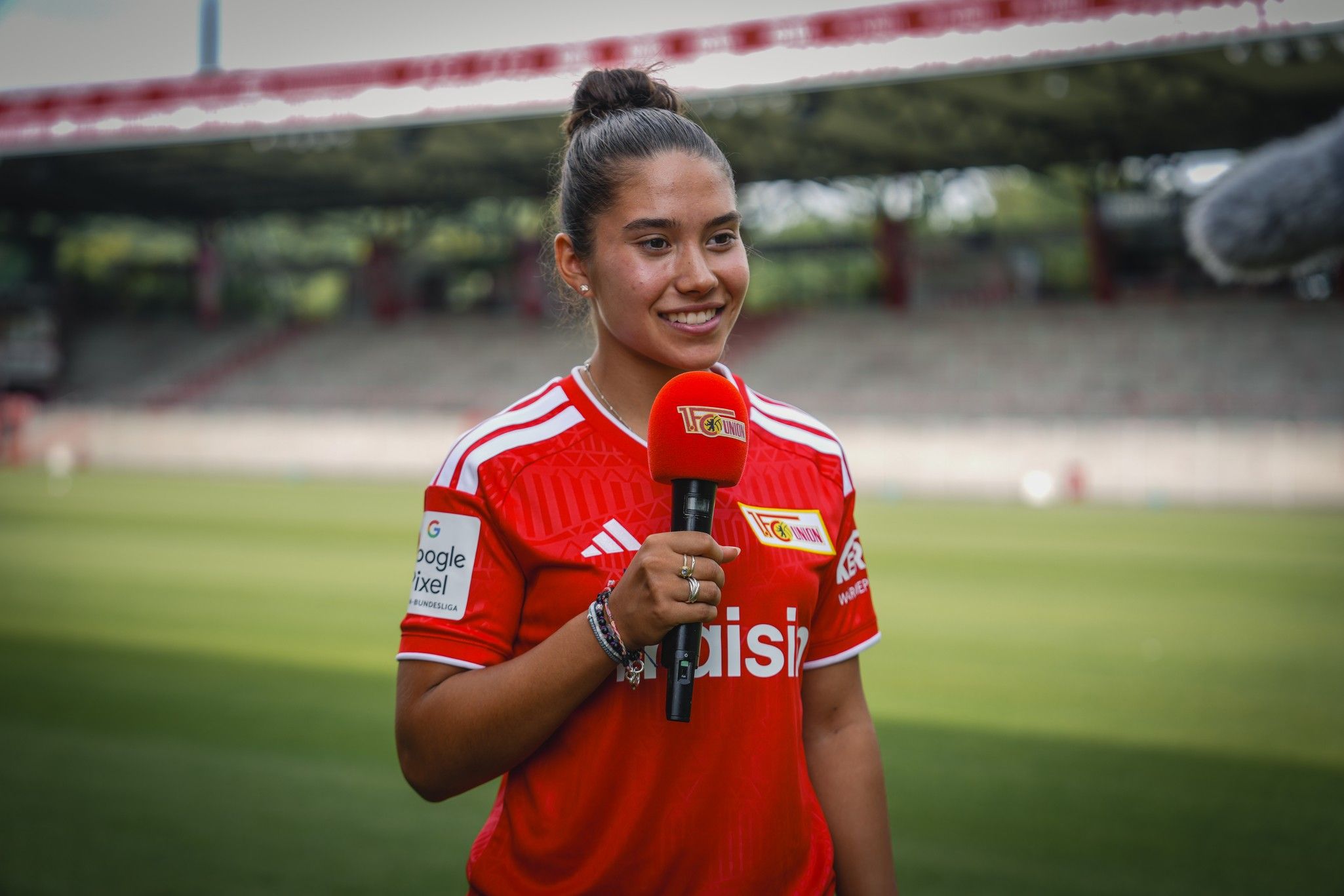Eine junge Frau trägt ein rotes Fußballtrikot und hält ein Mikrofon in einem Stadion. Sie lächelt und spricht in die Kamera.