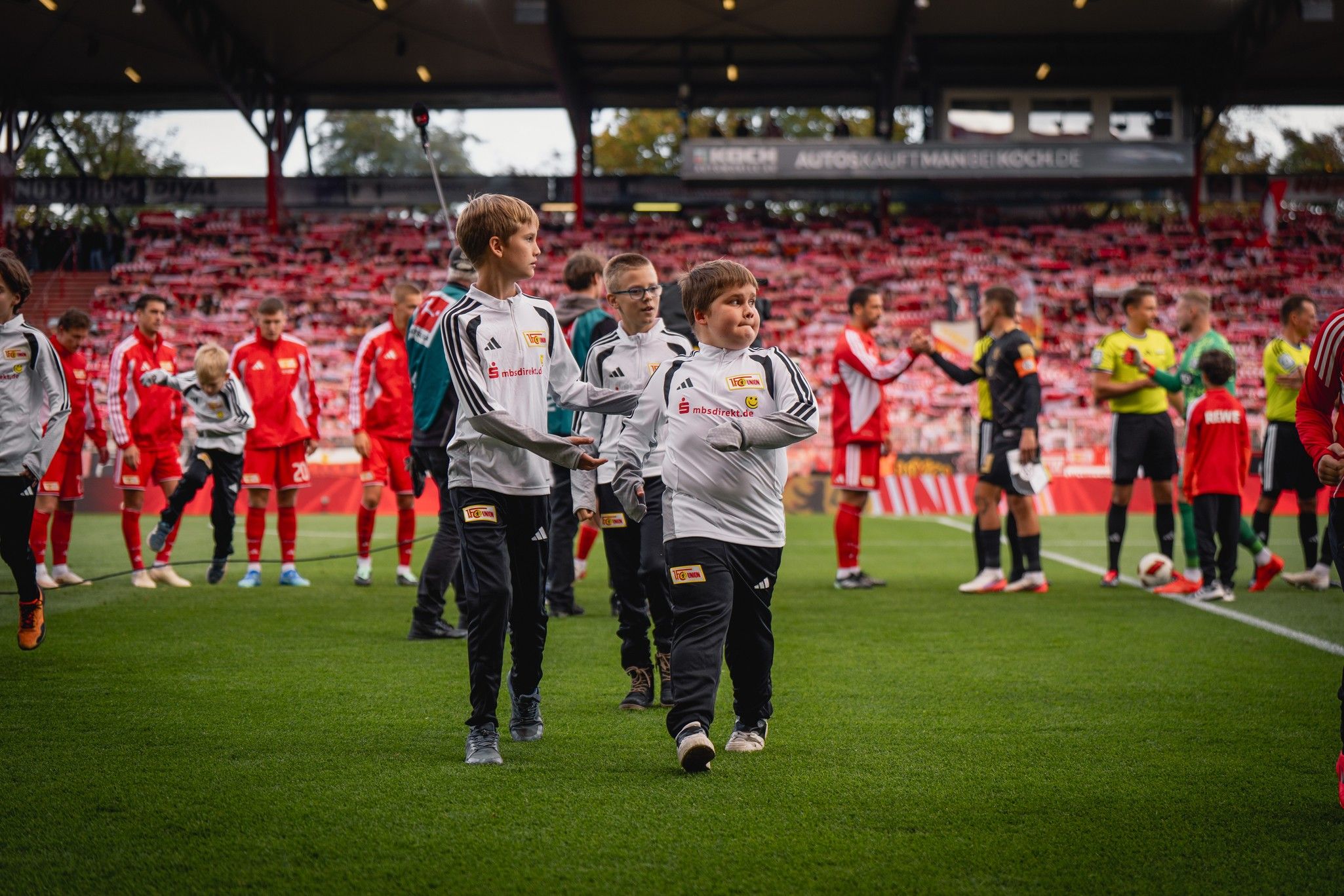 Kinder in Sportkleidung gehen auf das Spielfeld, begleitet von Spielern in roten Trikots, vor einem vollbesetzten Stadion.