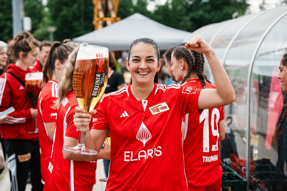 Eine lächelnde Frau in rotem Trikot hält ein großes Bierglas und zeigt ein Siegerpose. Im Hintergrund sind weitere Frauen in ähnlichen Trikots.