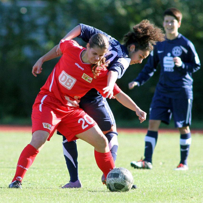Zwei Fußballspielerinnen kämpfen um den Ball auf einem Rasenplatz, während eine Zuschauerin im Hintergrund zuschaut.