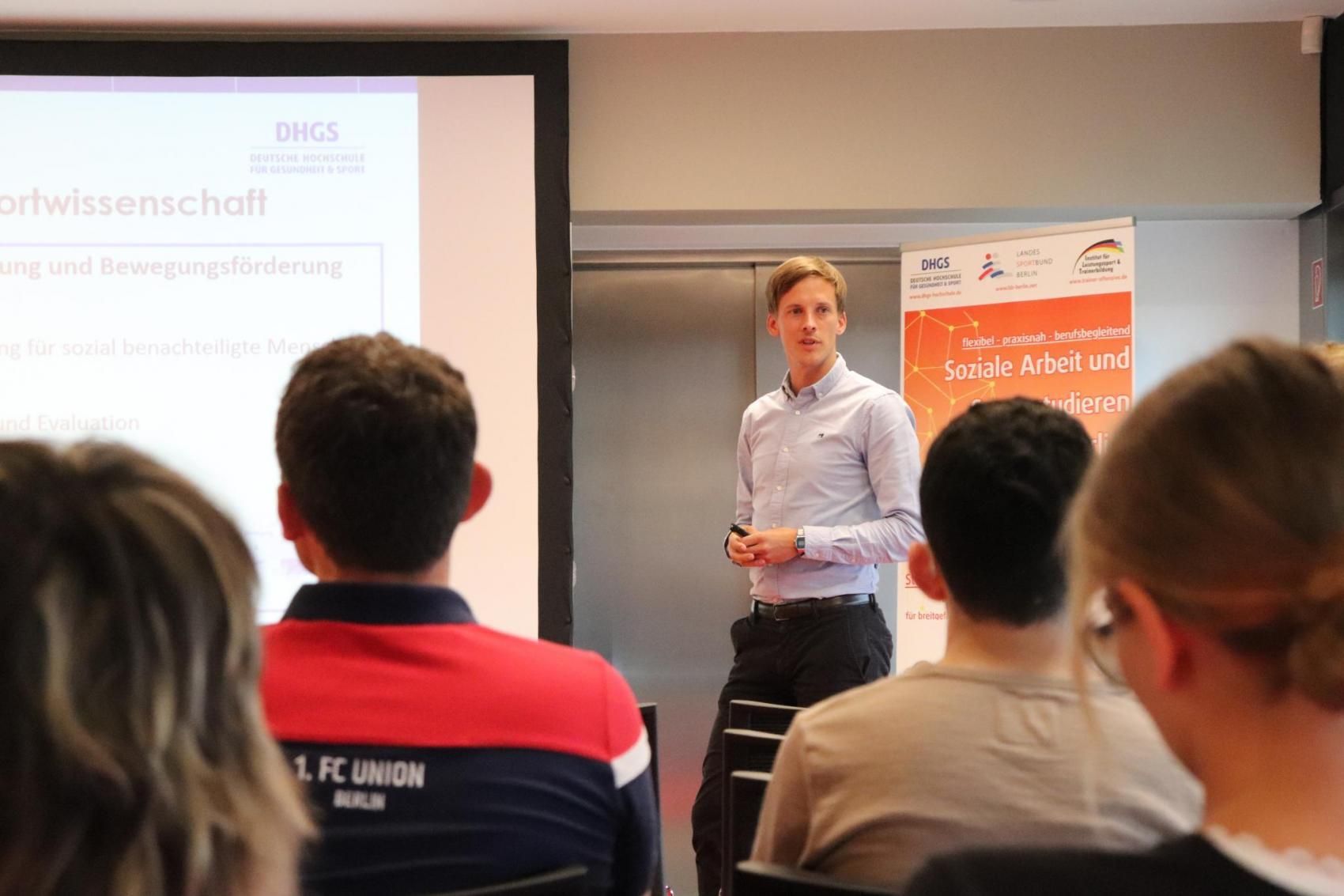 Ein Mann steht vor einem Publikum und präsentiert Informationen auf einer Folie, w ährend das Publikum ihm zuhört.