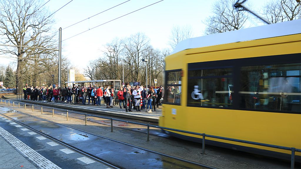 Menschen warten an einer Straßenbahnhaltestelle, während eine gelbe Straßenbahn vorbeifährt. Bäume im Hintergrund sind sichtbar.