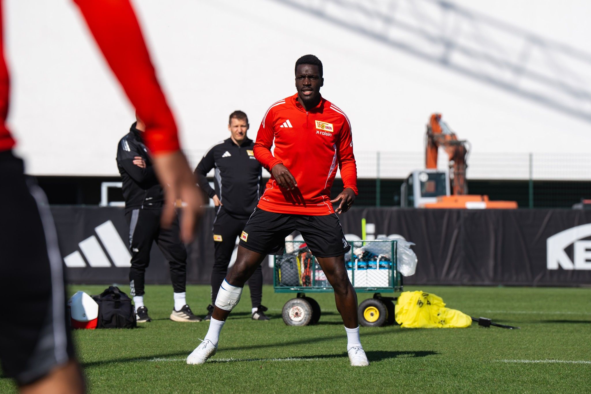 Ein Fußballspieler in rotem Trikot trainiert auf einem Platz, während andere Spieler und Mitarbeiter im Hintergrund zu sehen sind.