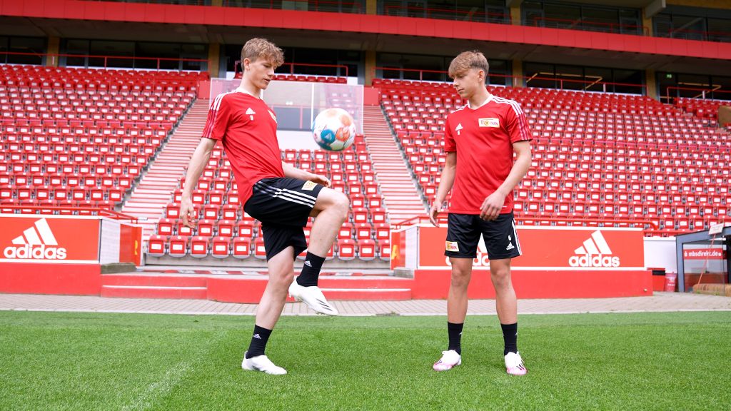 Zwei Jungen in roten Trikots jonglieren einen Fußball auf einem Stadionrasen mit roten Sitzplätzen im Hintergrund.