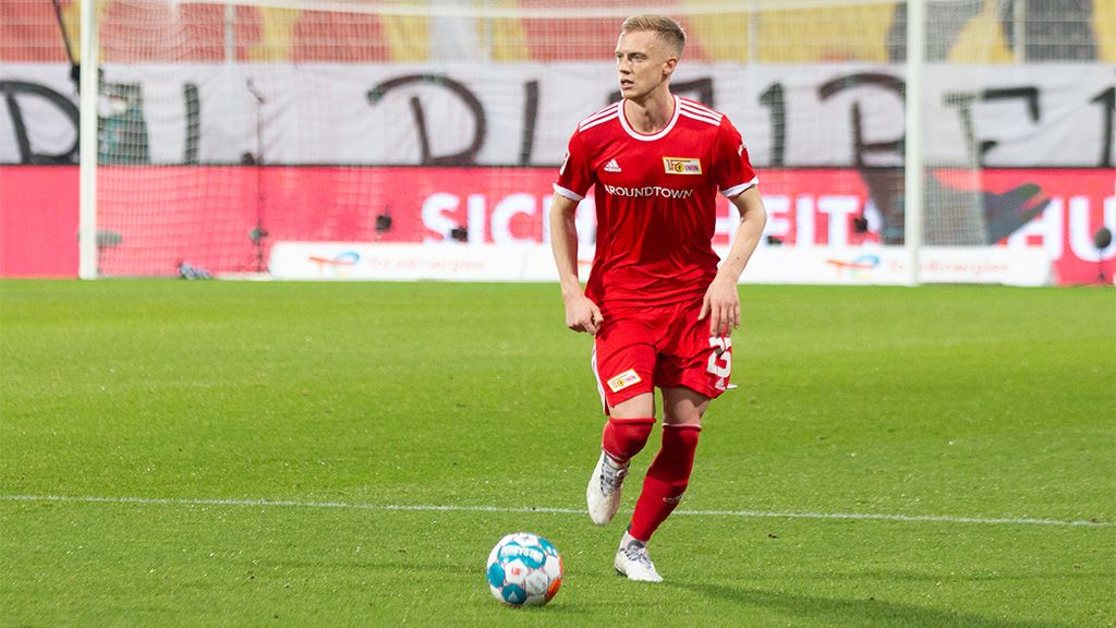 A soccer player in a red jersey is dribbling the ball on a soccer field. Banners are visible in the background.