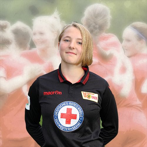 Ein Mädchen in einem schwarzen Fußballtrikot steht vor einer verschwommenen Gruppe von Mädchen in rot, die miteinander feiern.