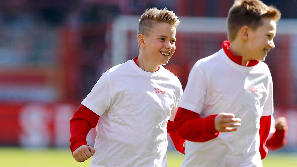 Zwei Kinder in weißen T-Shirts mit roten Ärmeln rennen fröhlich auf einem Sportplatz.