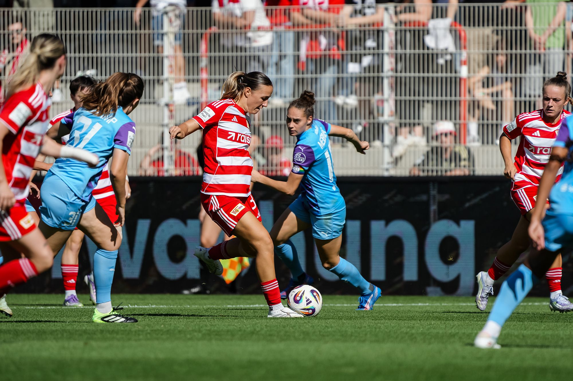 Zwei Frauenfußballspielerinnen kämpfen um den Ball auf einem Spielfeld, während andere Spielerinnen im Hintergrund zuschauen.