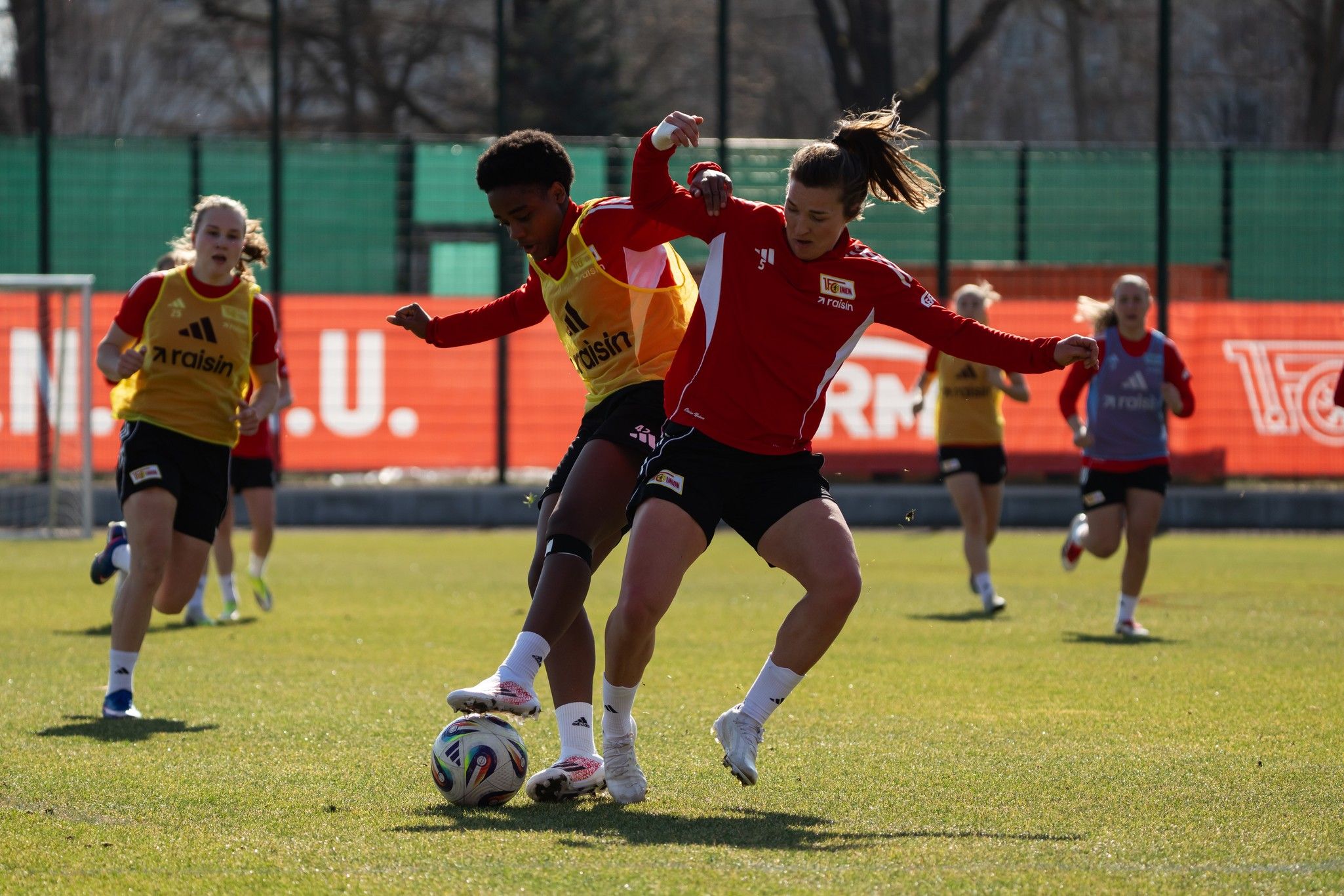 Zwei Fußballspielerinnen im Wettkampf um den Ball während eines Trainings auf einem grünen Platz, im Hintergrund weitere Spielerinnen.
