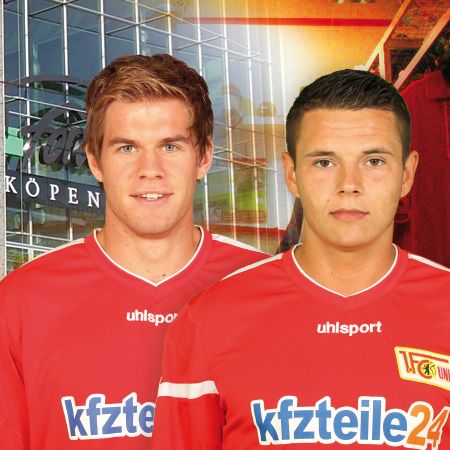 Zwei Fußballspieler in roten Trikots mit Sponsorenschriftzug, vor einem verschwommenen Stadionhintergrund.