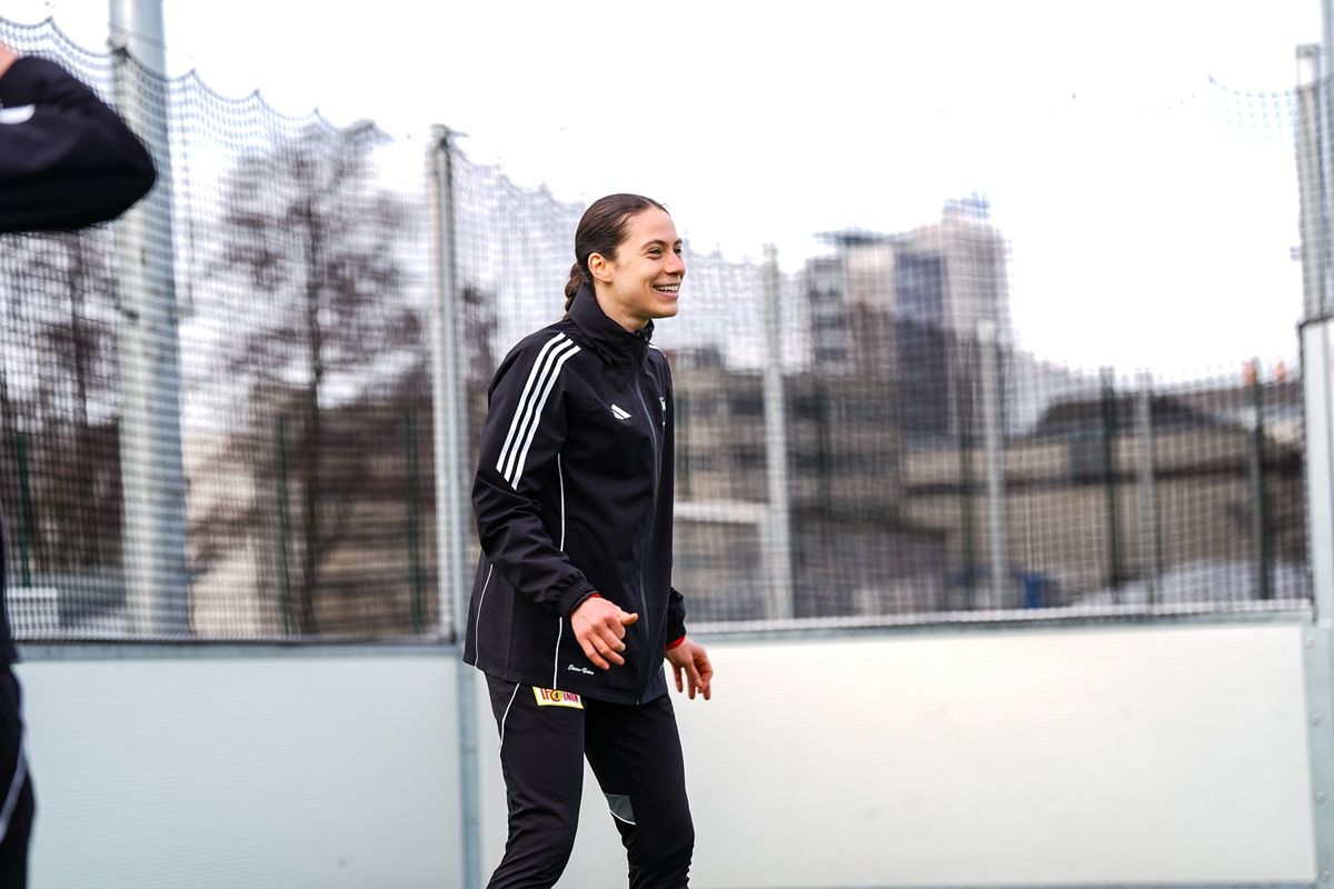 Eine Sportlerin in Trainingskleidung lächelt während des Trainings auf einem Fußballplatz. Im Hintergrund sind moderne Gebäude sichtbar.