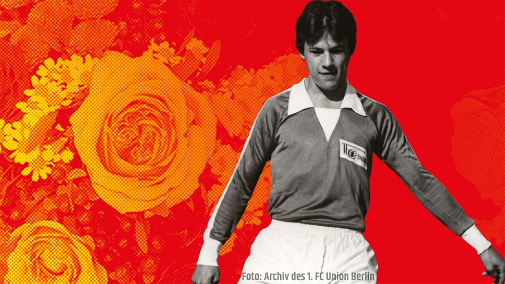 Ein Fußballspieler in einem grauen Trikot steht vor einem roten Hintergrund mit orangefarbenen Rosen.