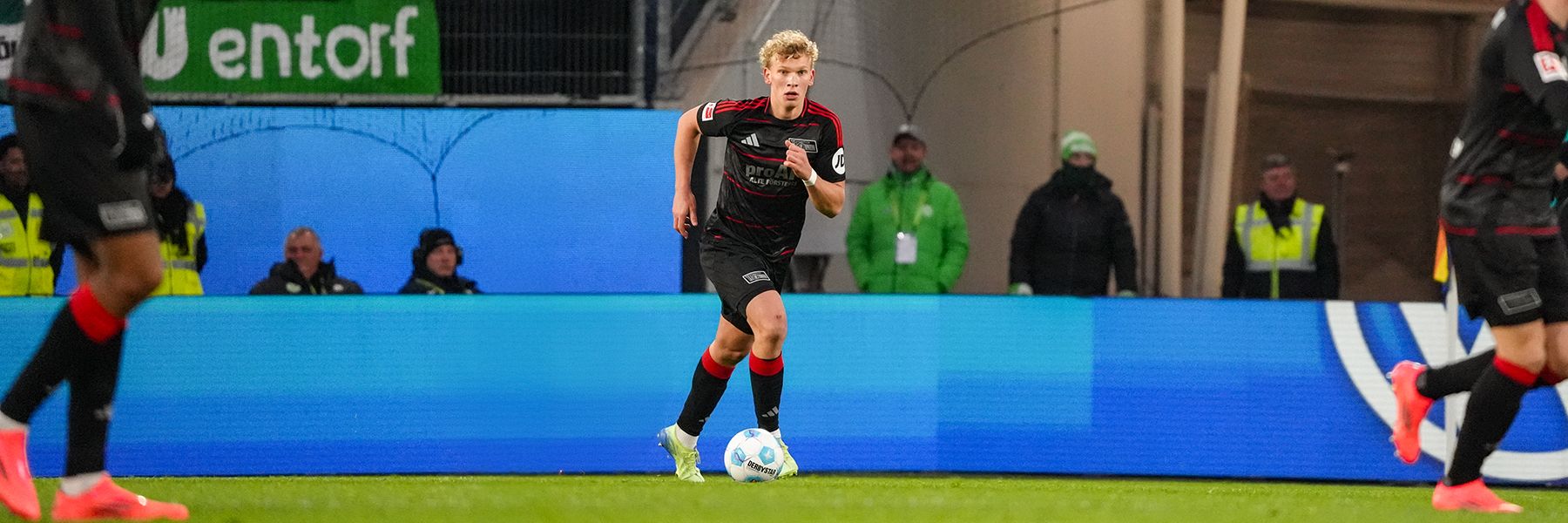 A soccer player in a black and red uniform dribbles the ball on the field, surrounded by other players and spectators.