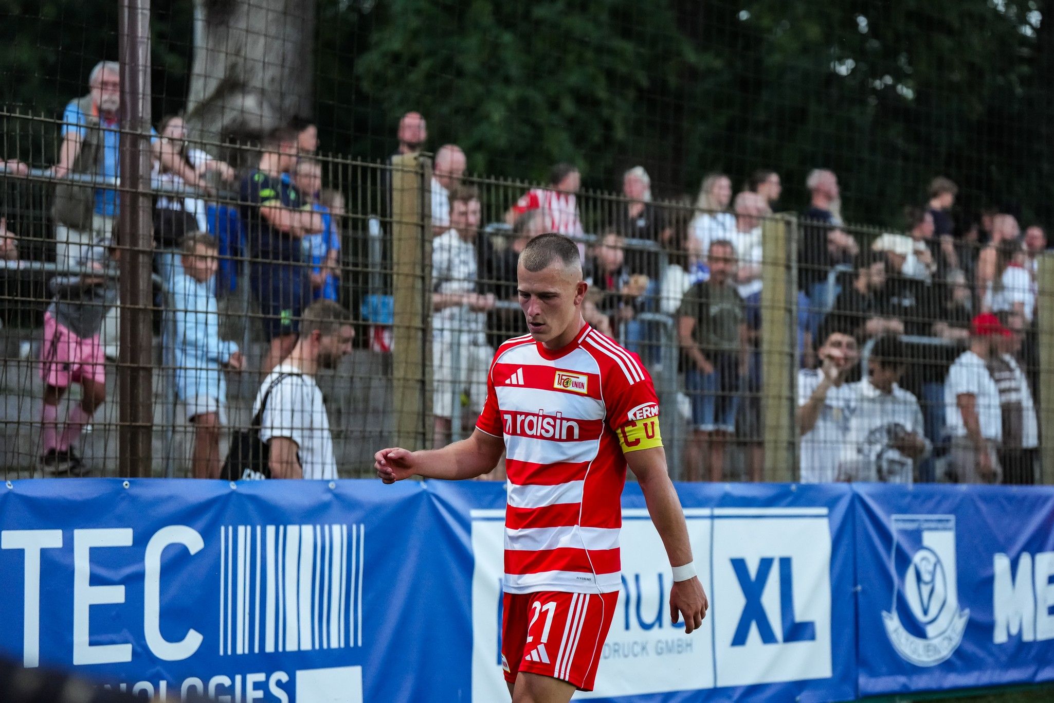 Ein Fußballspieler in einem rot-weißen Trikot geht niedergeschlagen am Spielfeldrand entlang, während Fans im Hintergrund zuschauen.