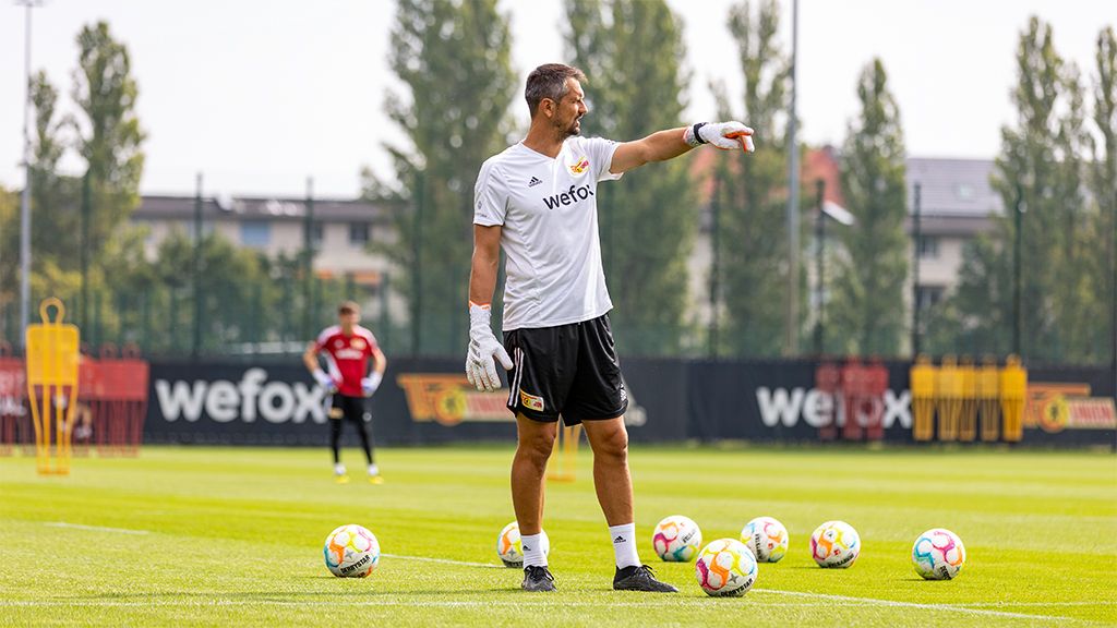 The goalkeeper trains on the field and gives instructions to a teammate while several soccer balls lie around him.