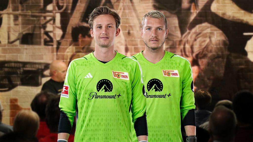 Zwei Torhüter in grünen Trikots stehen nebeneinander vor einer verschwommenen Kulisse.