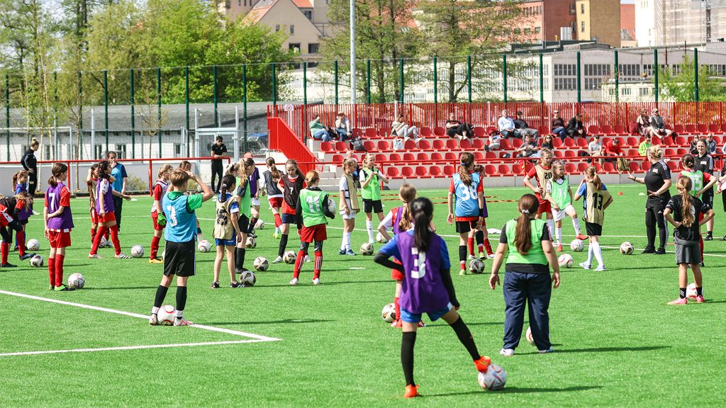 Mädchen unterschiedlichen Alters bei einem Fußballtraining auf einem Kunstrasenplatz. Zuschauer sitzen auf einer Tribüne im Hintergrund.