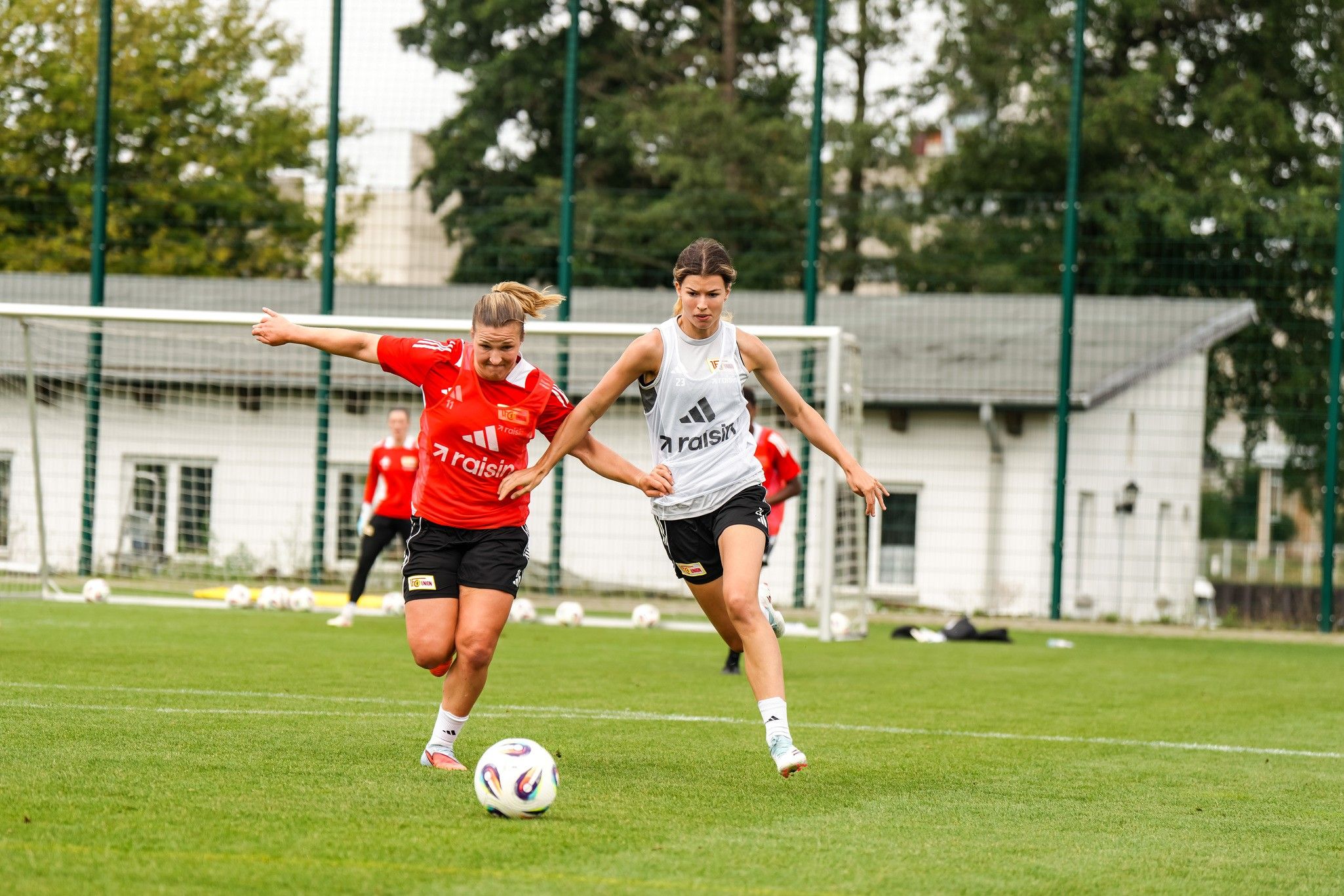 Zwei Fußballspielerinnen in einem Wettkampf auf einem Sportplatz mit Toren im Hintergrund.