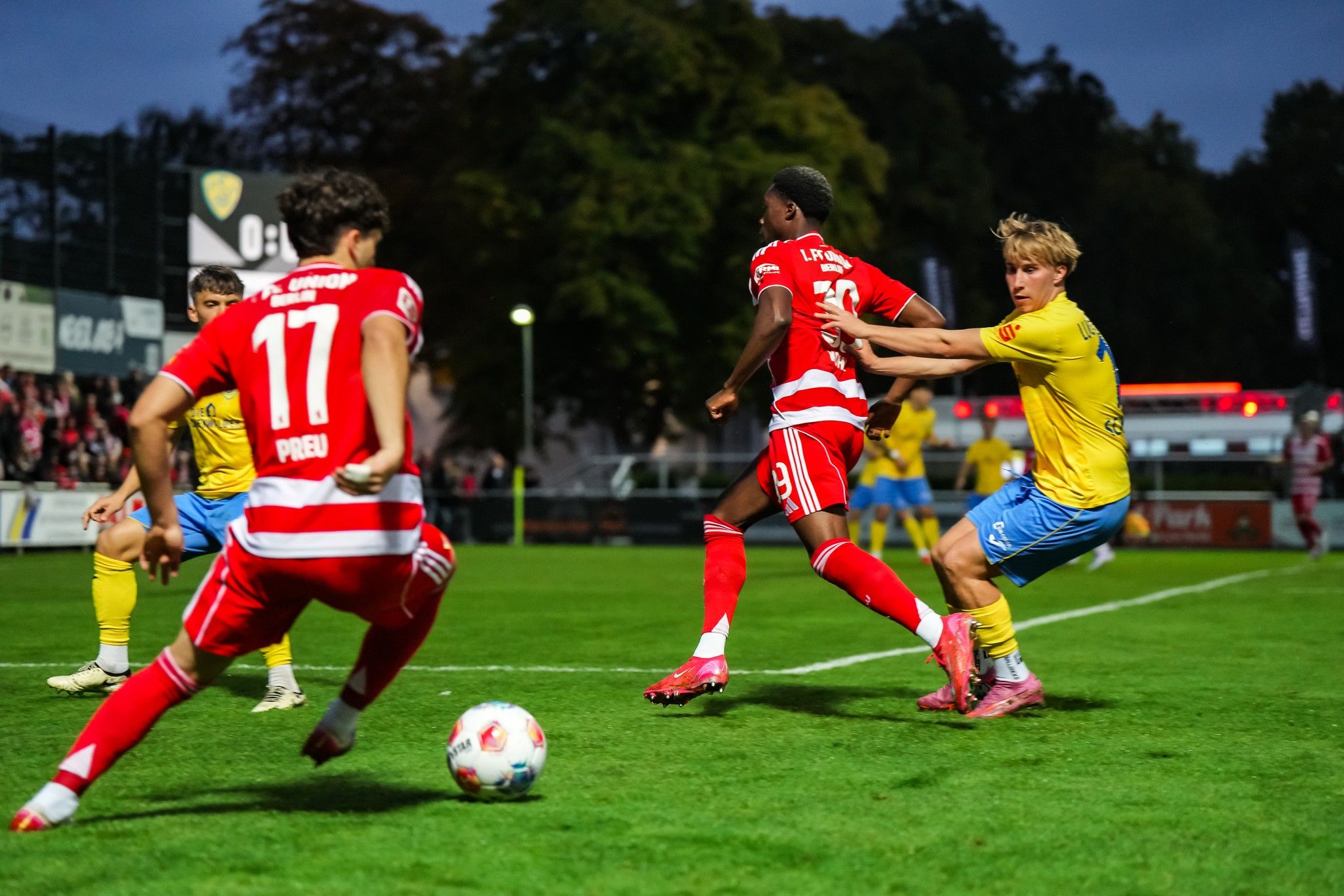 Zwei Fußballspieler im roten Trikot dribbeln, während ein Spieler im blauen Trikot versucht, den Ball zu erobern.