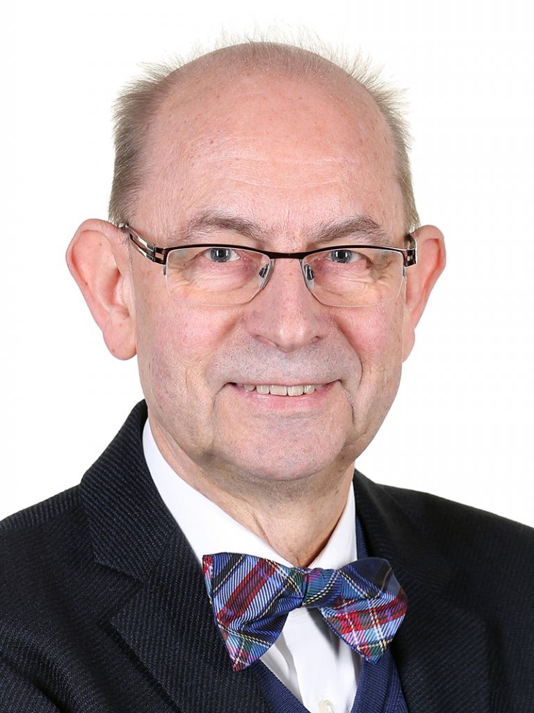 Porträt eines älteren Mannes mit Brille, Krawatte im Schottenmuster und einem schwarzen Anzug. Hintergrund ist hell und einfarbig.