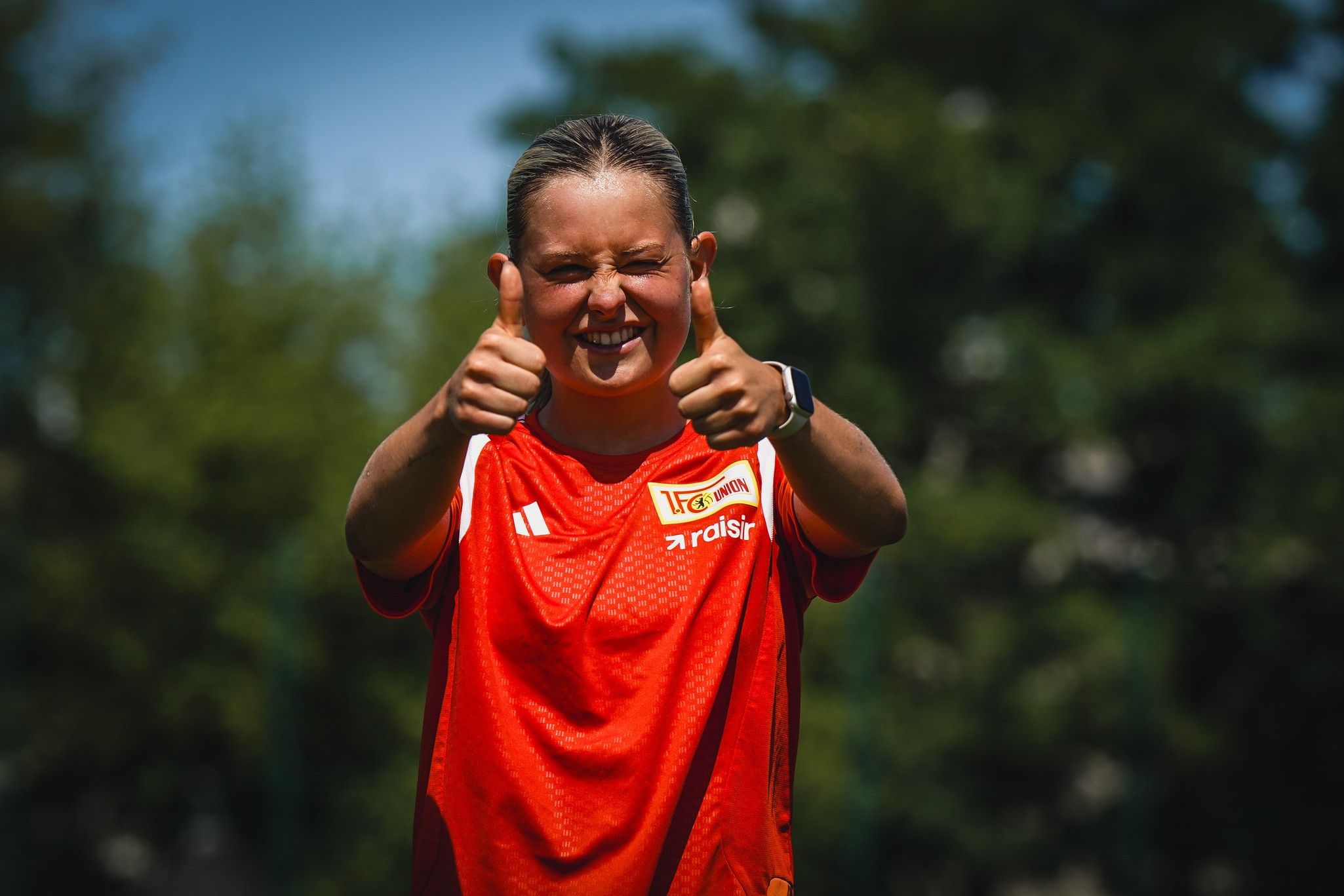 Eine junge Frau in einem roten Fußballtrikot zeigt mit beiden Händen Daumen nach oben und lächelt fröhlich.