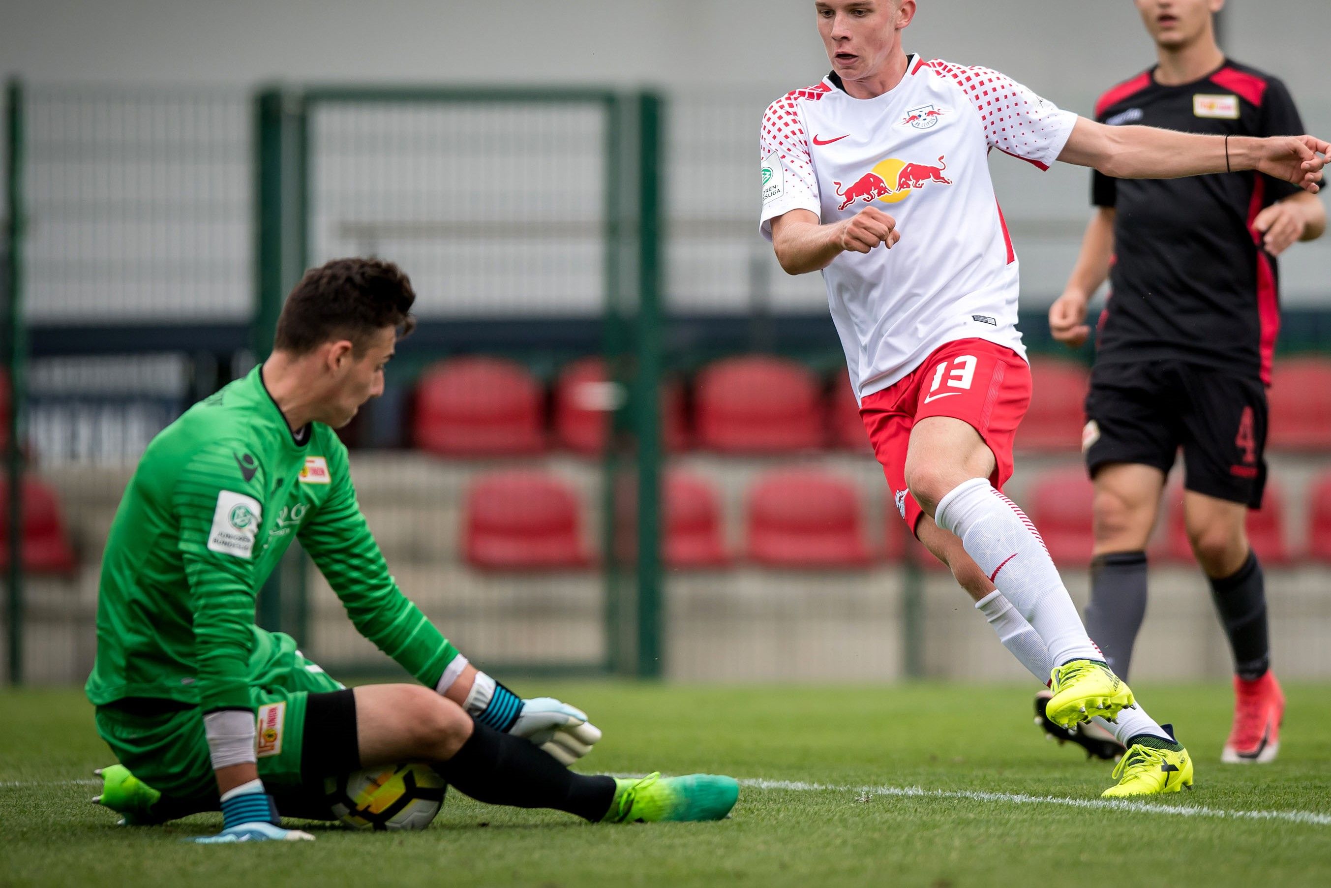 Ein Fußballspieler dribbelt am Torwart vorbei, während dieser am Boden sitzt. Die Szene spielt auf einem Fußballfeld.