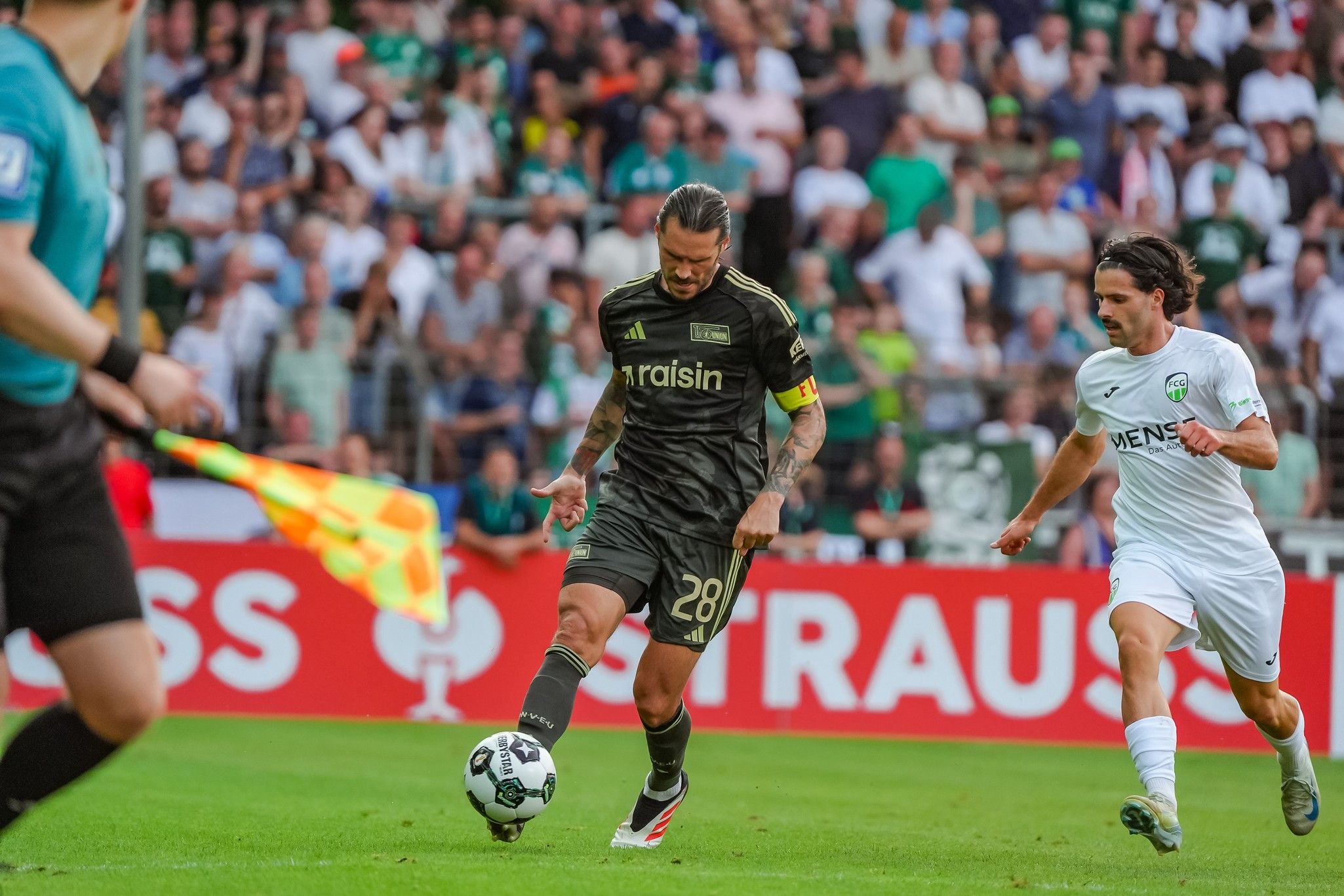 Ein Fußballspieler in dunkler Ausrüstung dribbelt den Ball, während ein Gegner in weißer Ausrüstung ihn verfolgt. Zuschauer im Hintergrund.