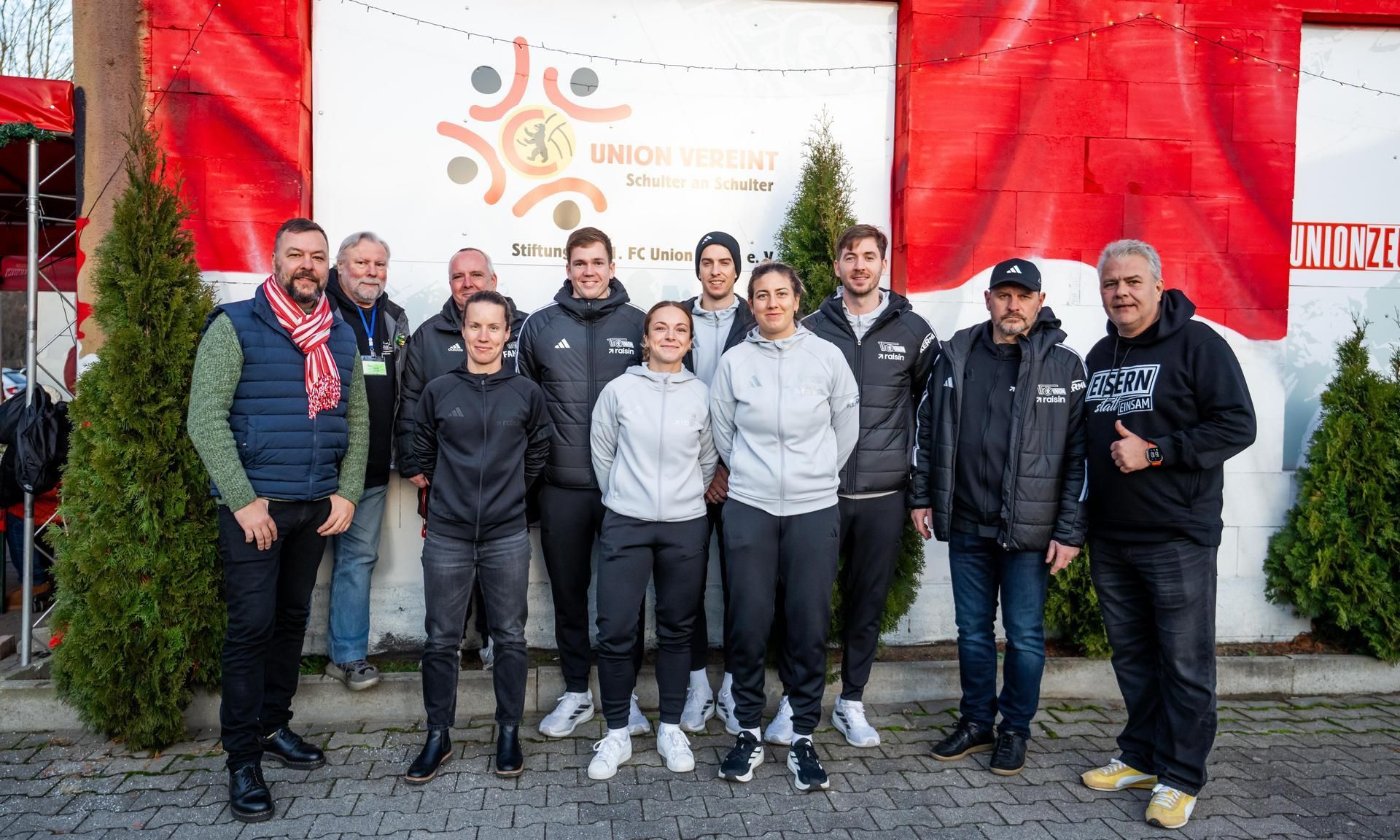 Eine Gruppe von Personen steht vor einer Wand mit dem Logo von "Union Verein". Sie tragen Freizeitkleidung und lächeln in die Kamera.