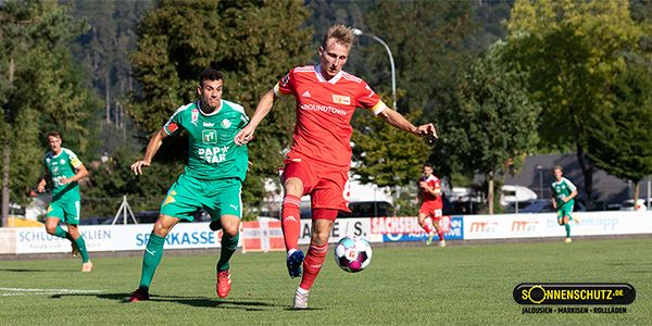 A soccer player in a red jersey with a ball, being chased by a player in a green goalkeeper jersey. A sports field scene under a blue sky.