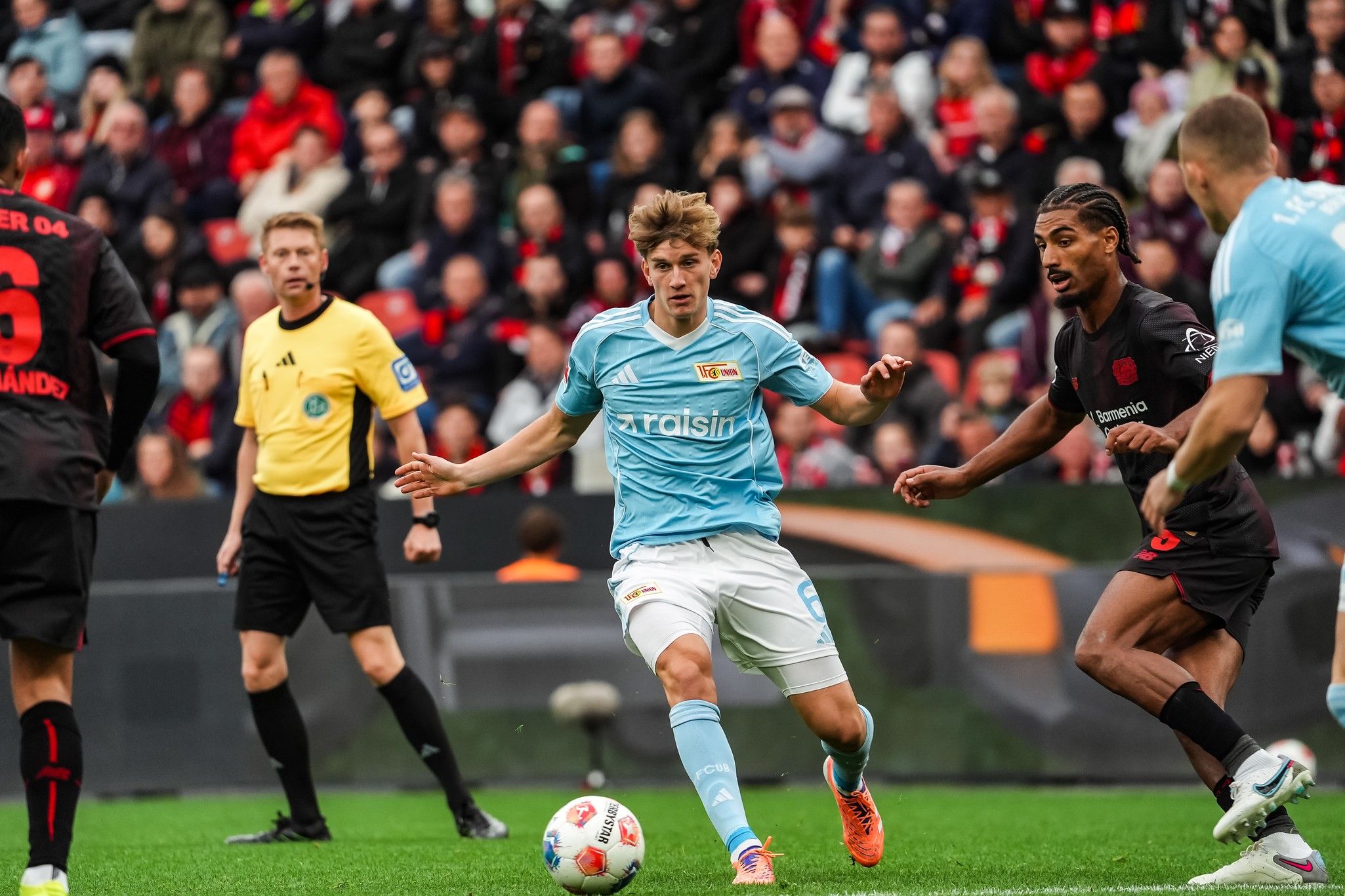 Spielszene eines Fußballspiels mit Spielern in blauen und schwarzen Trikots, einem Schiedsrichter im Hintergrund und einer Zuschauertribüne.