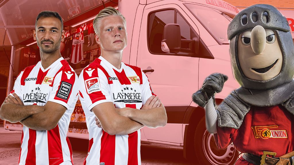 Zwei Fußballspieler im Heimtrikot mit roten und weißen Streifen stehen stolz nebeneinander, während ein Maskottchen in Rüstung lächelt.