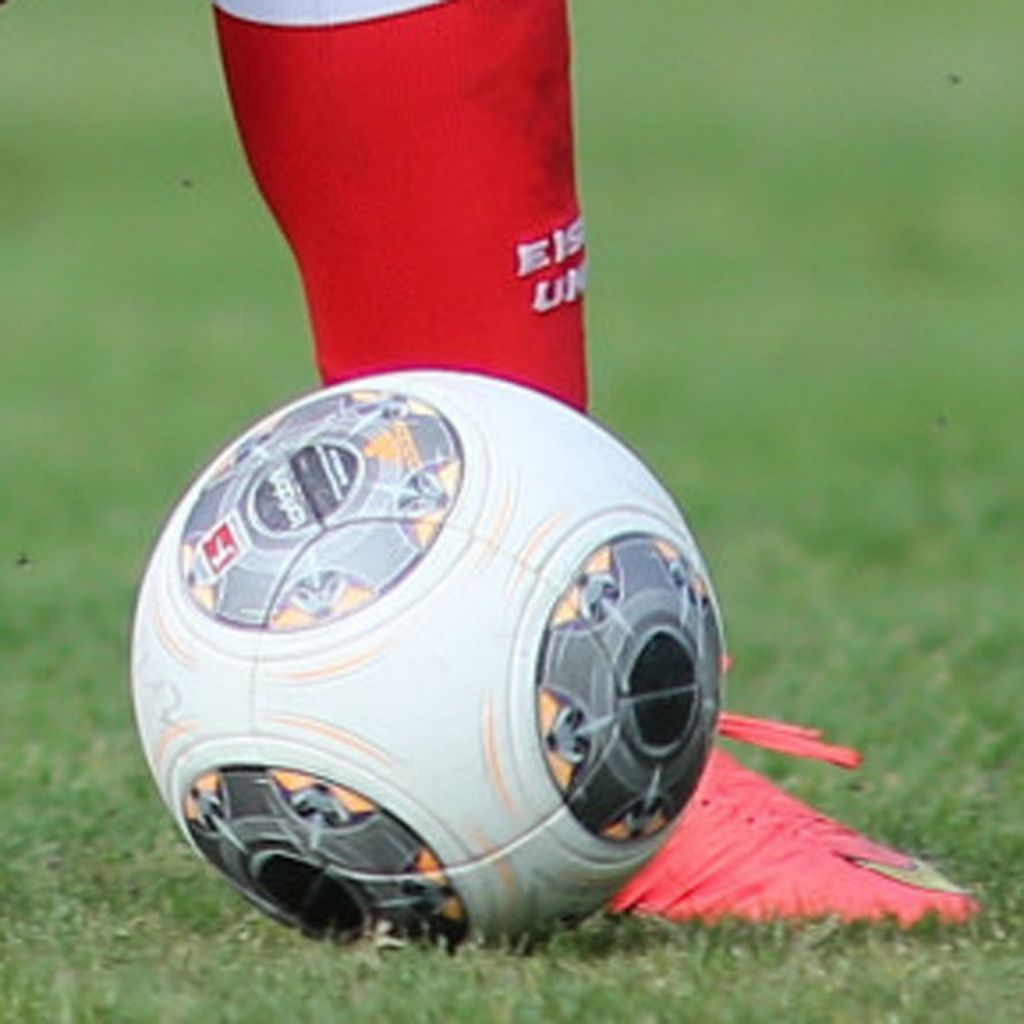 Ein Fußball liegt auf dem Rasen, während ein Spieler mit roter Socke und Schuhen in Fußballausrüstung daneben steht.