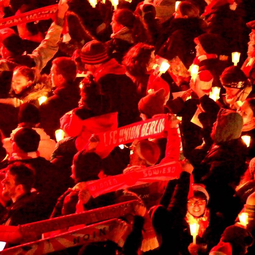 Menschenmenge mit Fackeln und Schals, die einen Fußballverein unterstützen, stehen in einem festlichen, roten Licht.