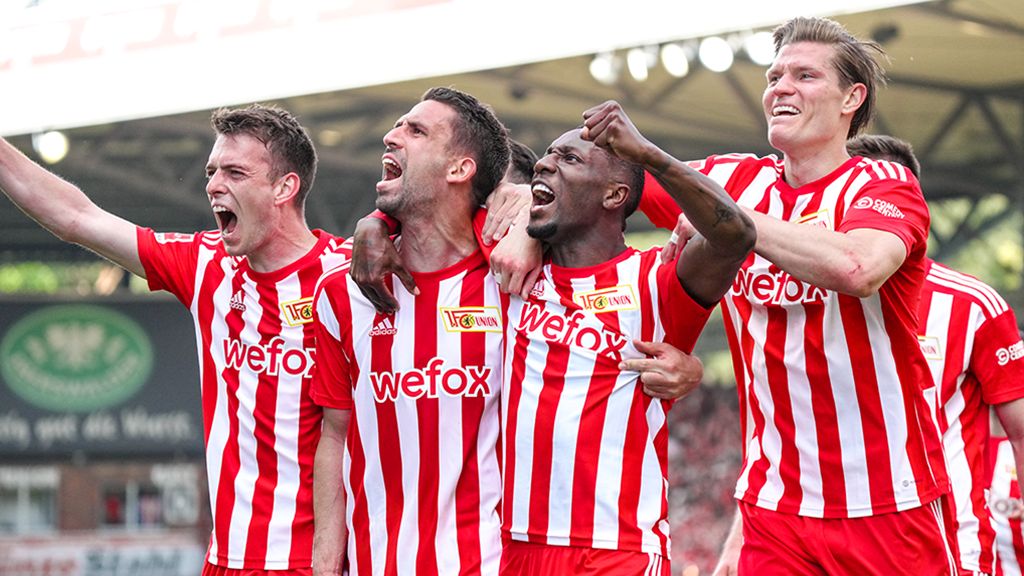 Vier Fußballspieler tragen rote und weiße Trikots. Sie feiern auf dem Spielfeld.