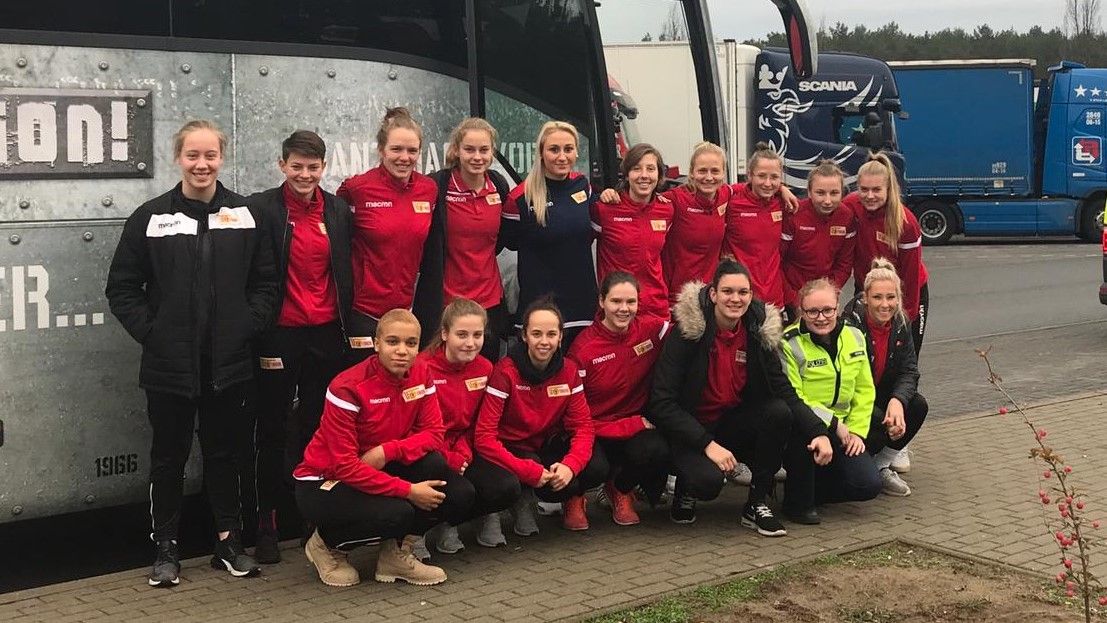 Gruppe von zwölf Sportlerinnen in roten Trainingsanzügen vor einem Bus, lächeln und posieren zusammen für das Foto.