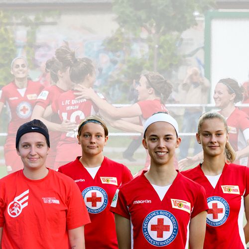Vier junge Frauen in Roter Kreuz-Trikots stehen zusammen, während im Hintergrund eine feiernde Fußballmannschaft zu sehen ist.