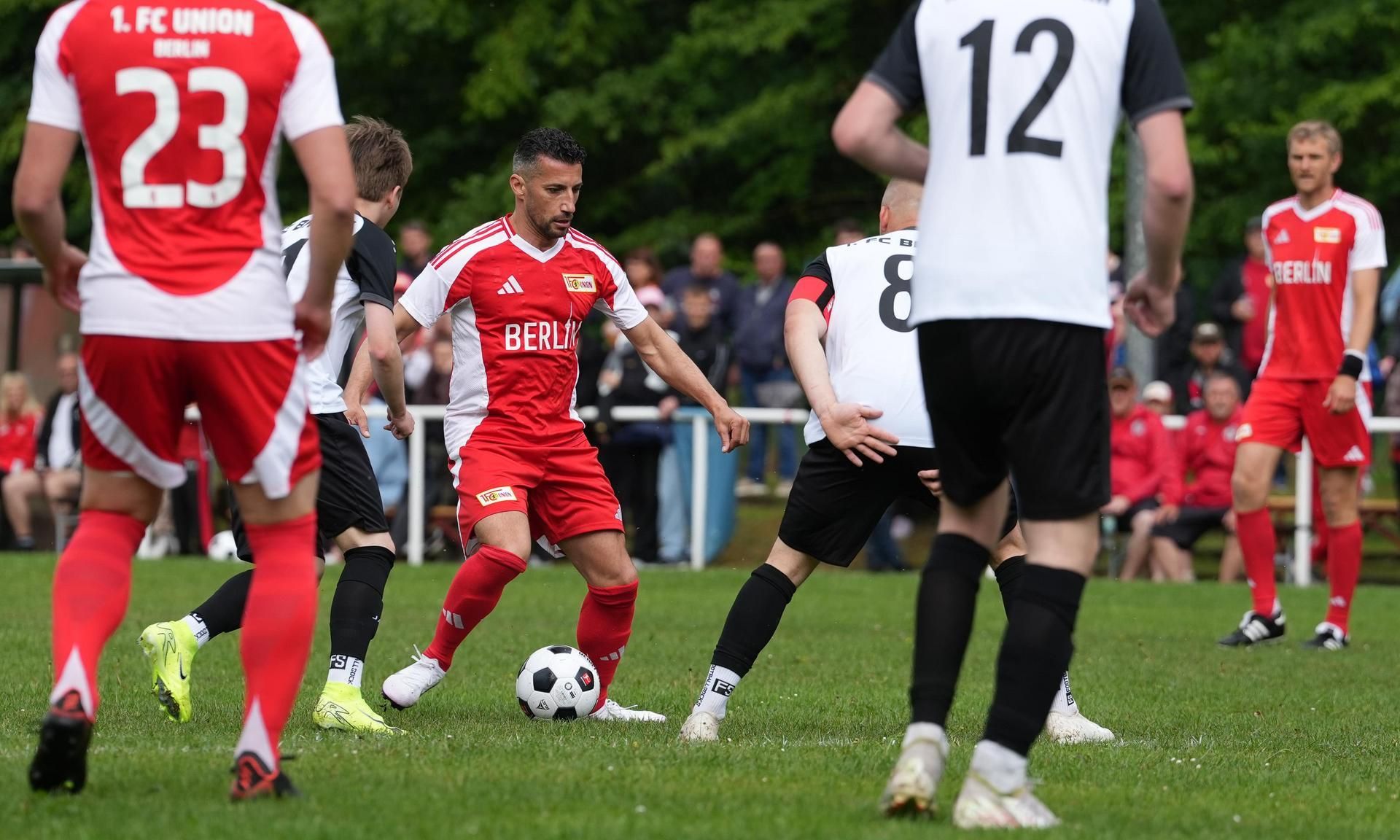 Ein Fußballspieler in roter Uniform dribbelt den Ball zwischen zwei Spielern in schwarz-weißen Trikots während eines Spiels auf einem grünen Platz.