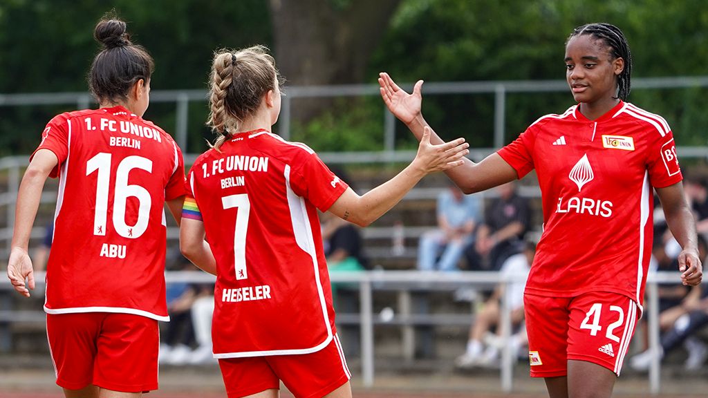 Zwei Fußballspielerinnen des 1. FC Union Berlin in roten Trikots begrüßen eine dritte Spielerin auf dem Spielfeld.